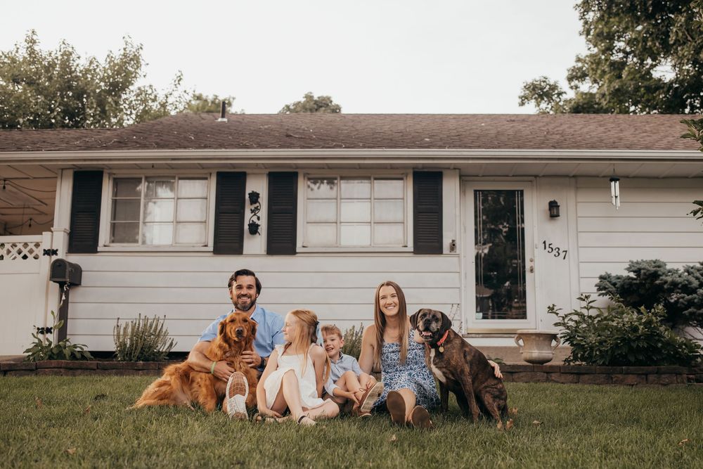 Family with two dogs sitting on grass in front of white ranch home for a portrait photo.