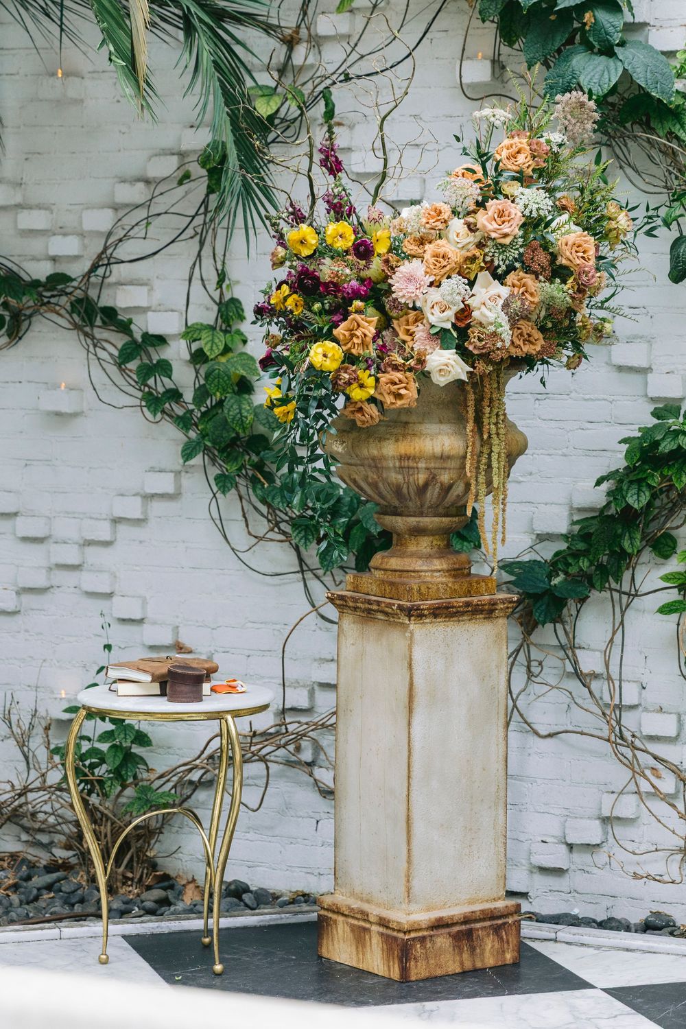 Elegant stone urn filled with colorful roses on a classical pedestal against a white brick wall with lush greenery.