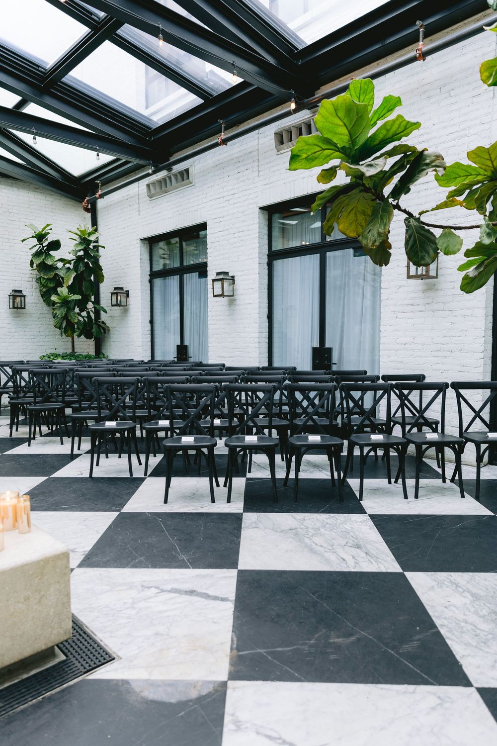 Elegant indoor venue with black and white checkered marble floors, dark cross-back chairs, and a glass ceiling with lush greenery.