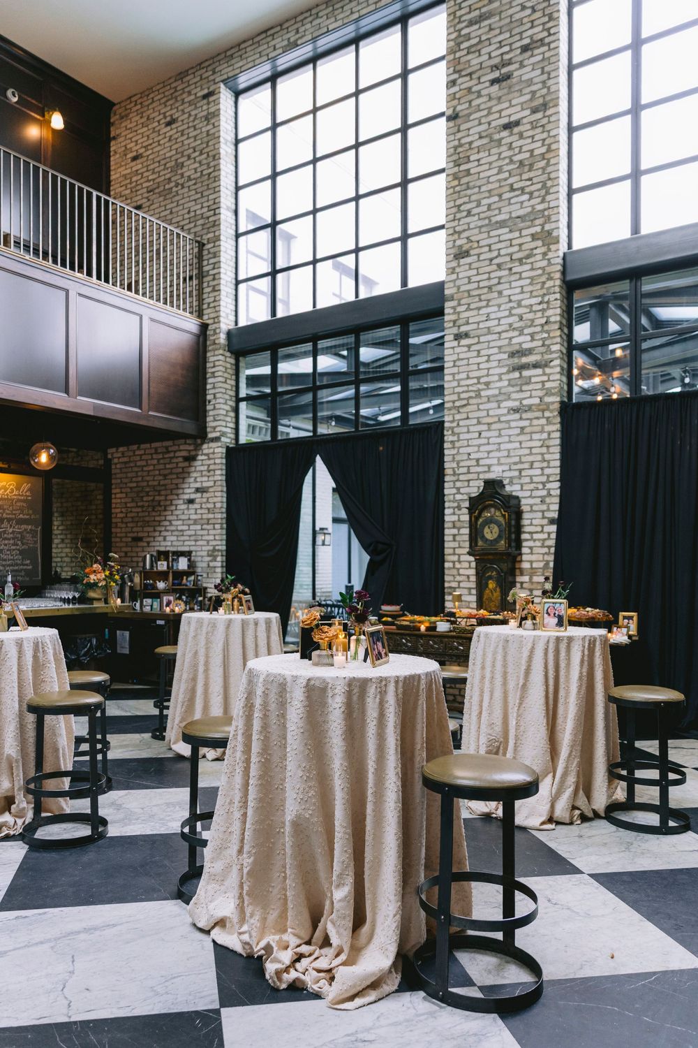 Industrial wedding venue with brick walls, high windows, checkered floors, and elegant draped cocktail tables with bar stools.
