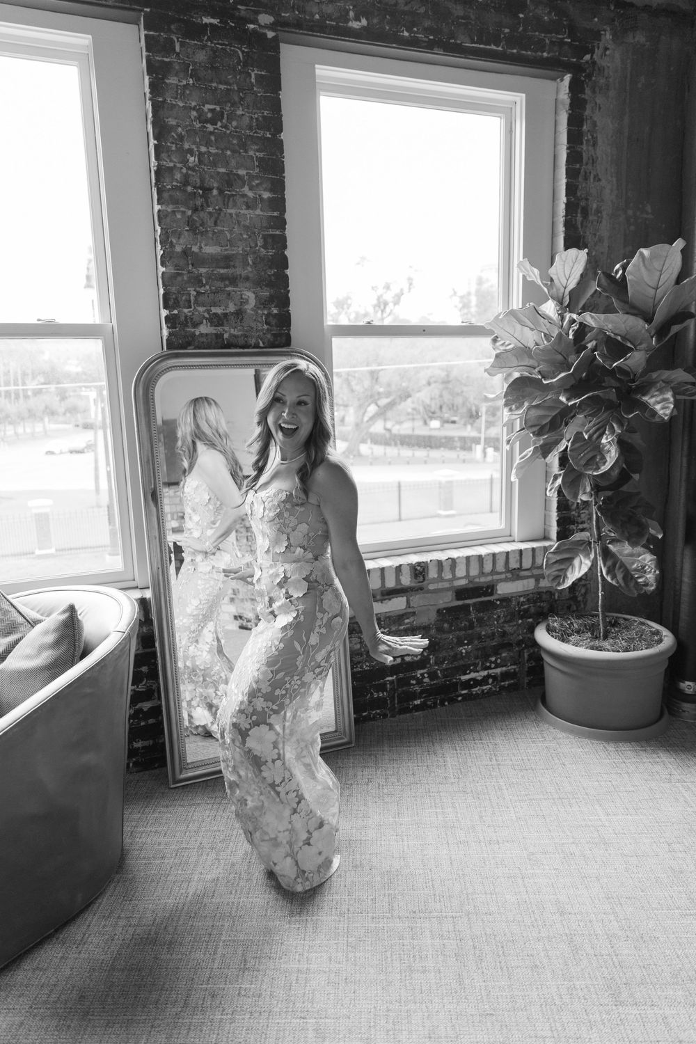 Black and white photo of a laughing bride in a floral gown standing beside a mirror in a room with brick walls.