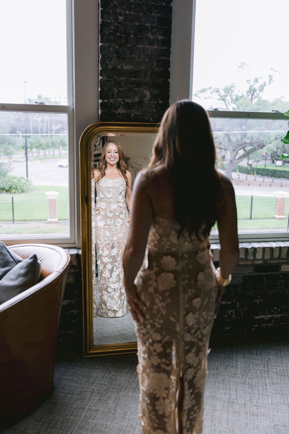 Bride in floral gown admires her reflection in a gold-framed mirror near a window in an elegant bridal suite.