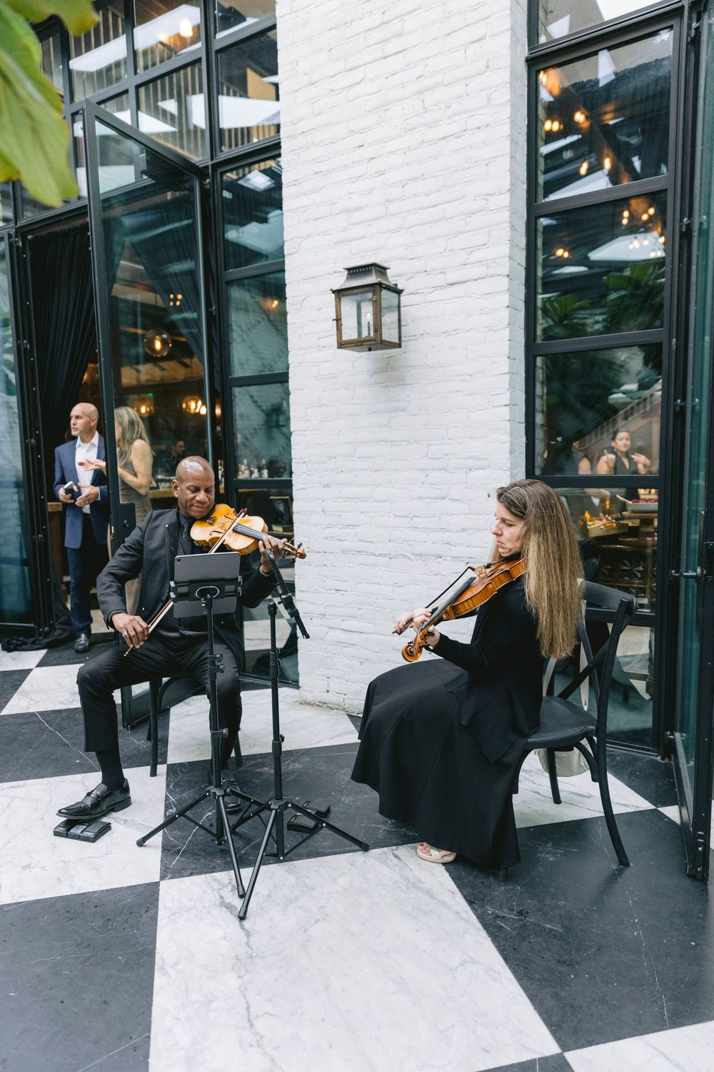 Two violinists perform outdoors on a checkered floor patio, dressed in black formal attire at an elegant event.