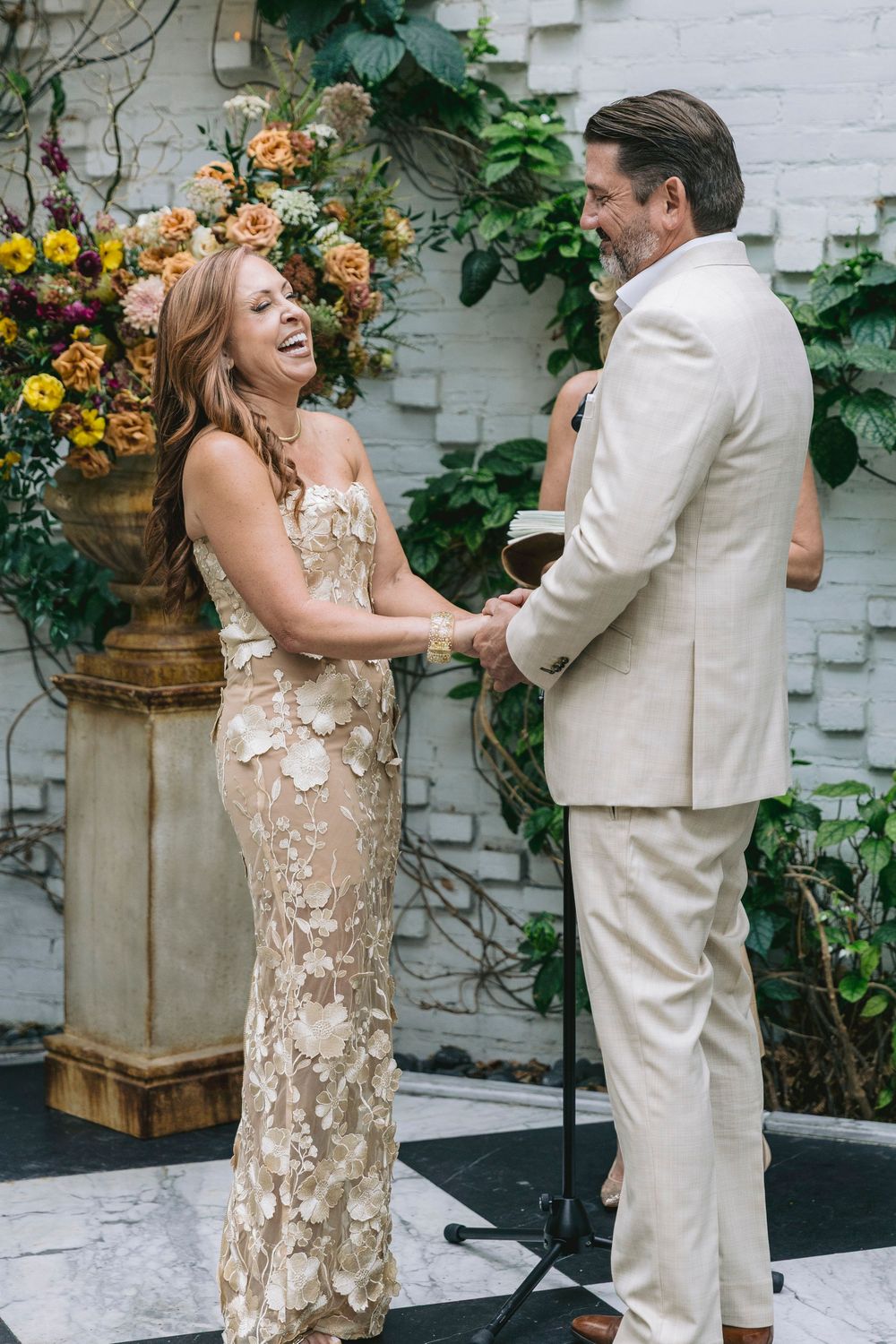 Joyful bride in gold floral gown laughing with groom in ivory suit during wedding ceremony with colorful floral backdrop.