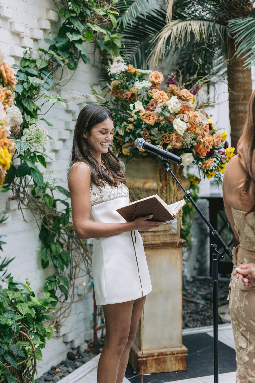 Woman in white dress reads from book at floral-decorated podium during outdoor wedding ceremony.