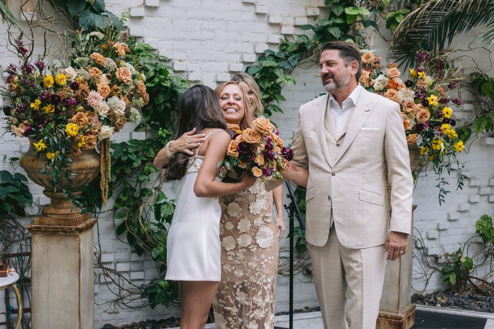 Bride in floral dress hugs guest at outdoor wedding ceremony with colorful floral arrangements on white brick wall.