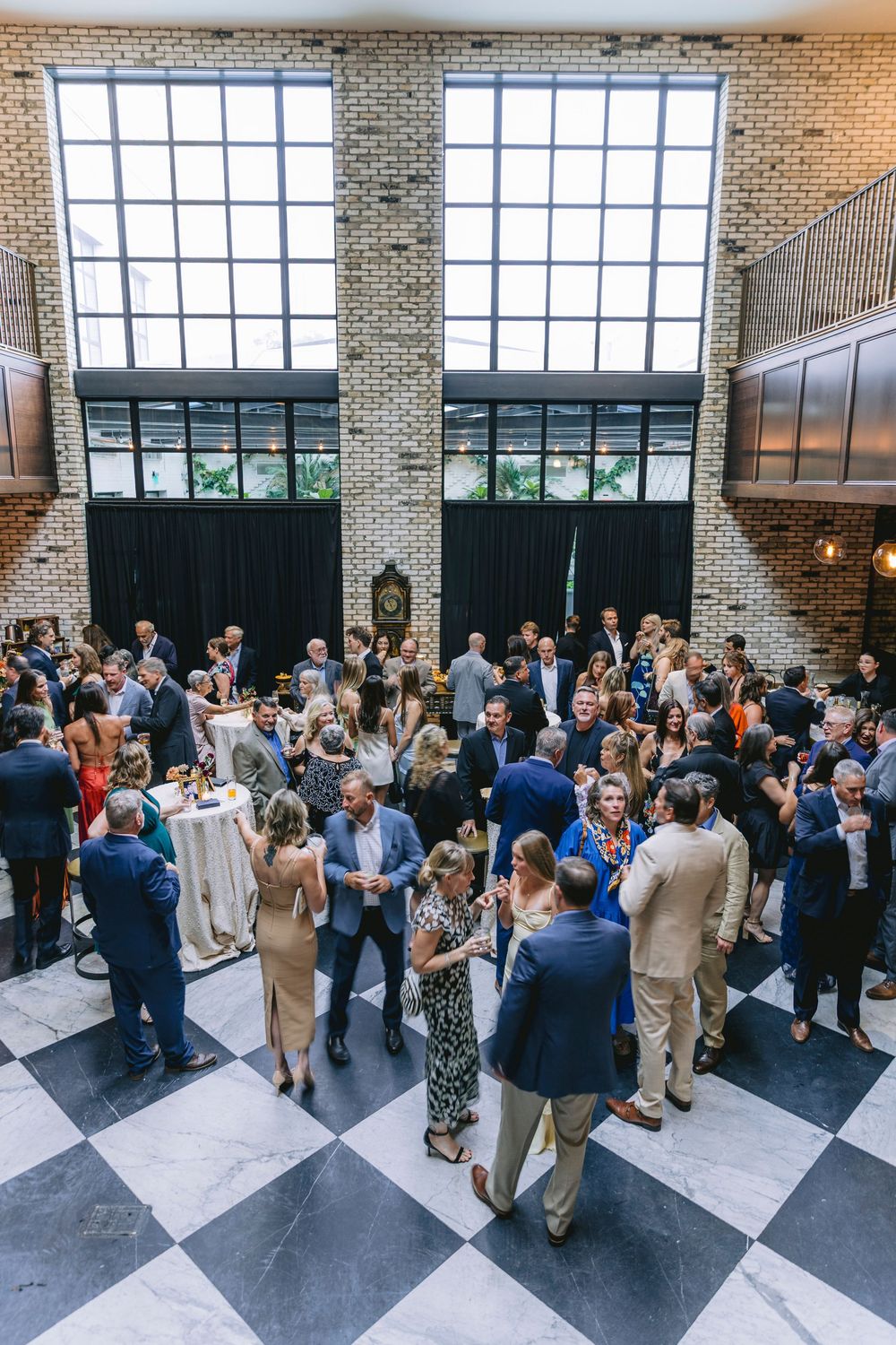 Aerial view of elegantly dressed guests mingling at an upscale event in a brick venue with checkered floors.