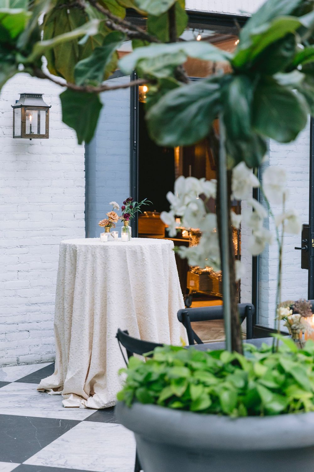 Elegant outdoor patio with a round table draped in linen, surrounded by lush greenery and white orchids near a warmly lit entrance.