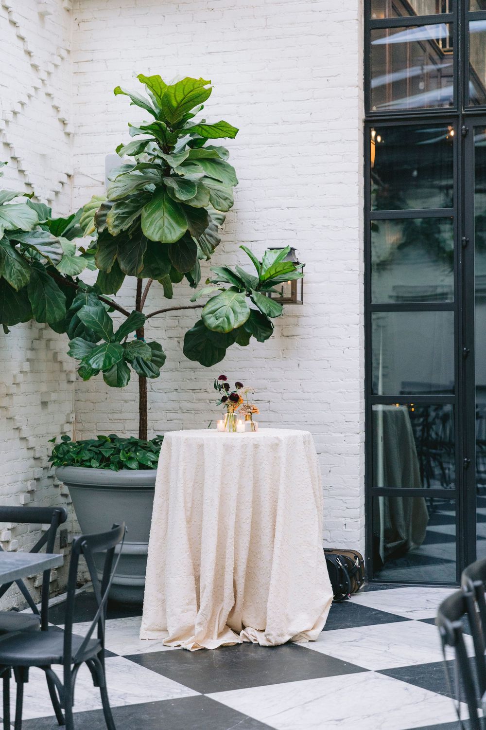 Outdoor cocktail hour setup with a cream sequin tablecloth, floral centerpiece, and large fiddle leaf fig tree against a white brick wall.