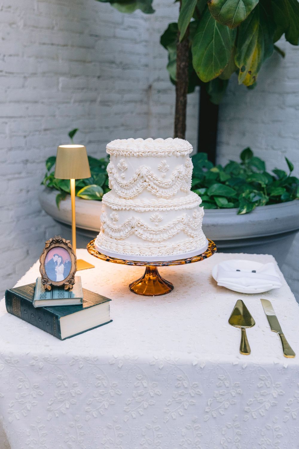 Two-tier white wedding cake with piped lace details on amber cake stand, displayed with gold serving set and small lamp.