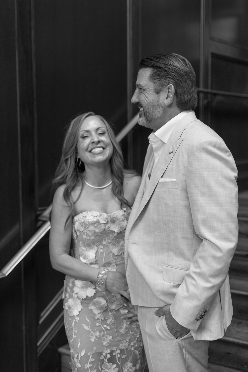 Black and white photo of a smiling couple in formal attire on stairs, woman in floral dress, man in light suit.