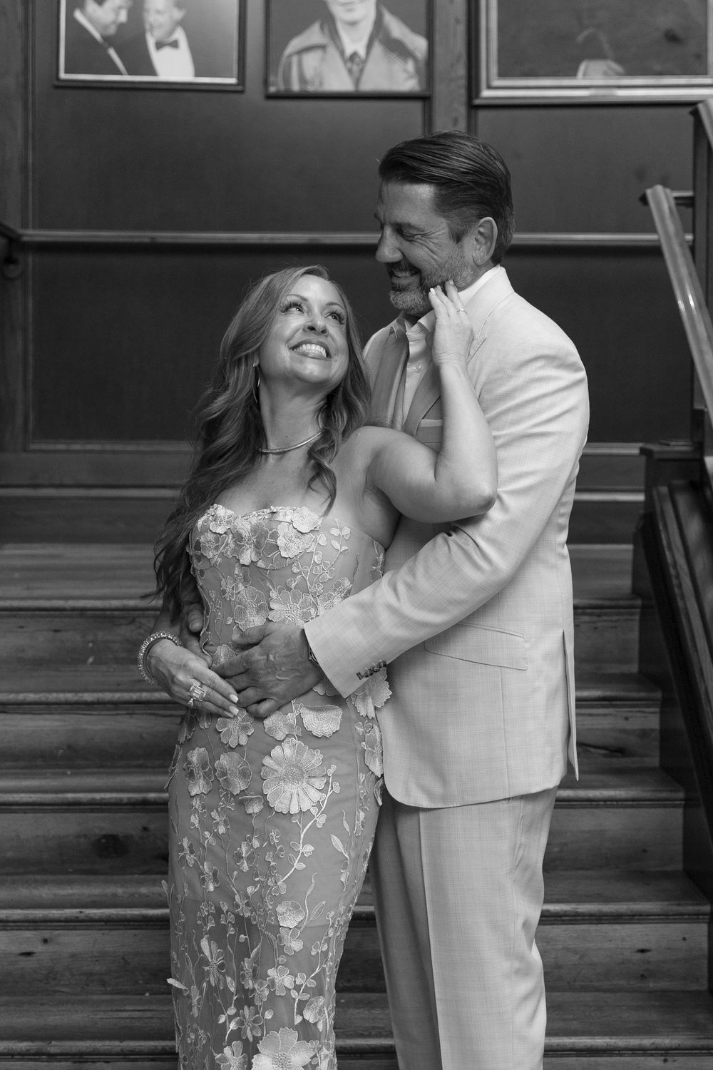 Black and white photo of a smiling couple in formal wear embracing on stairs, woman in floral gown, man in light suit.