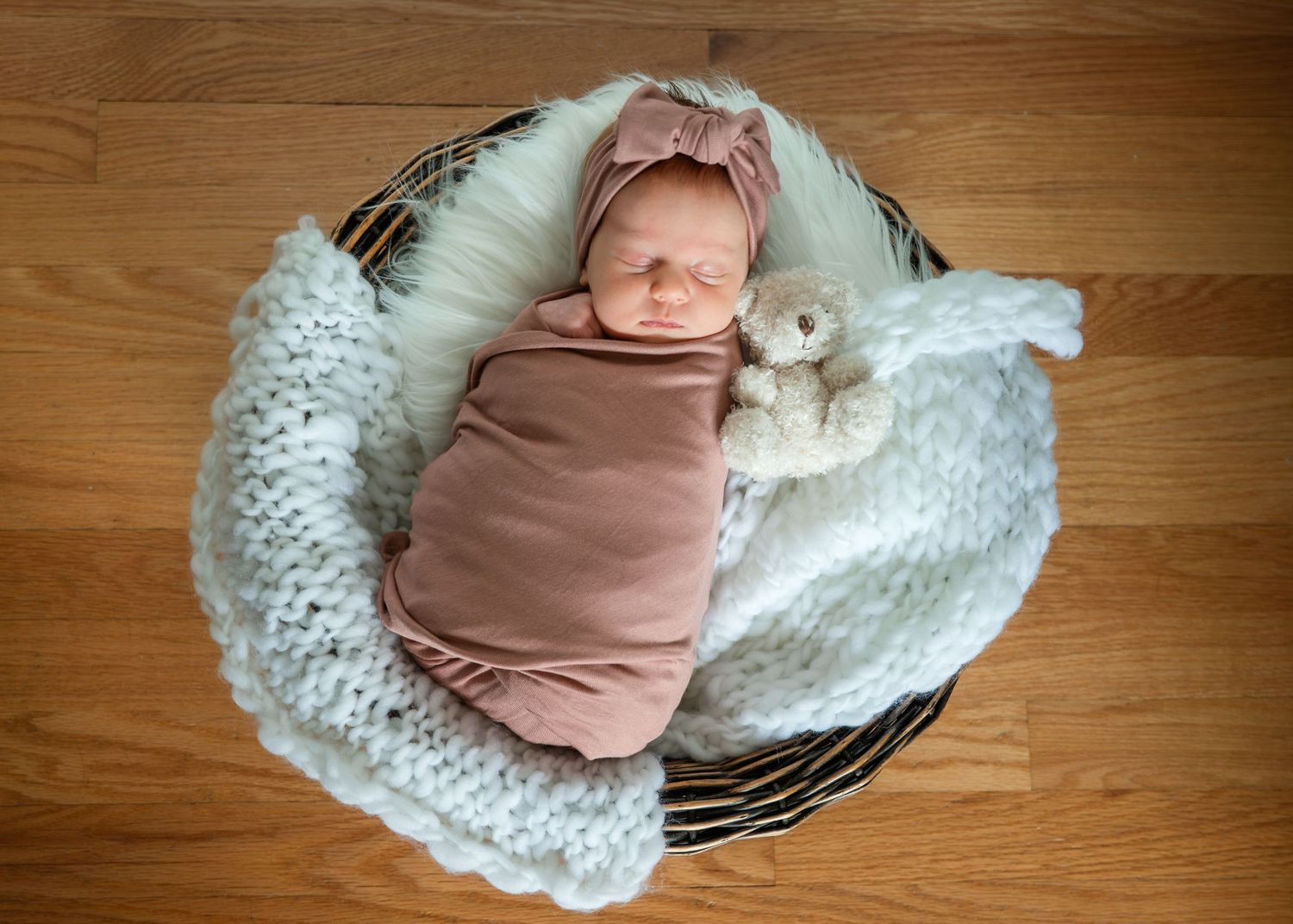 Newborn baby swaddled in pink wrap sleeping in a basket with a white knit blanket and small teddy bear.