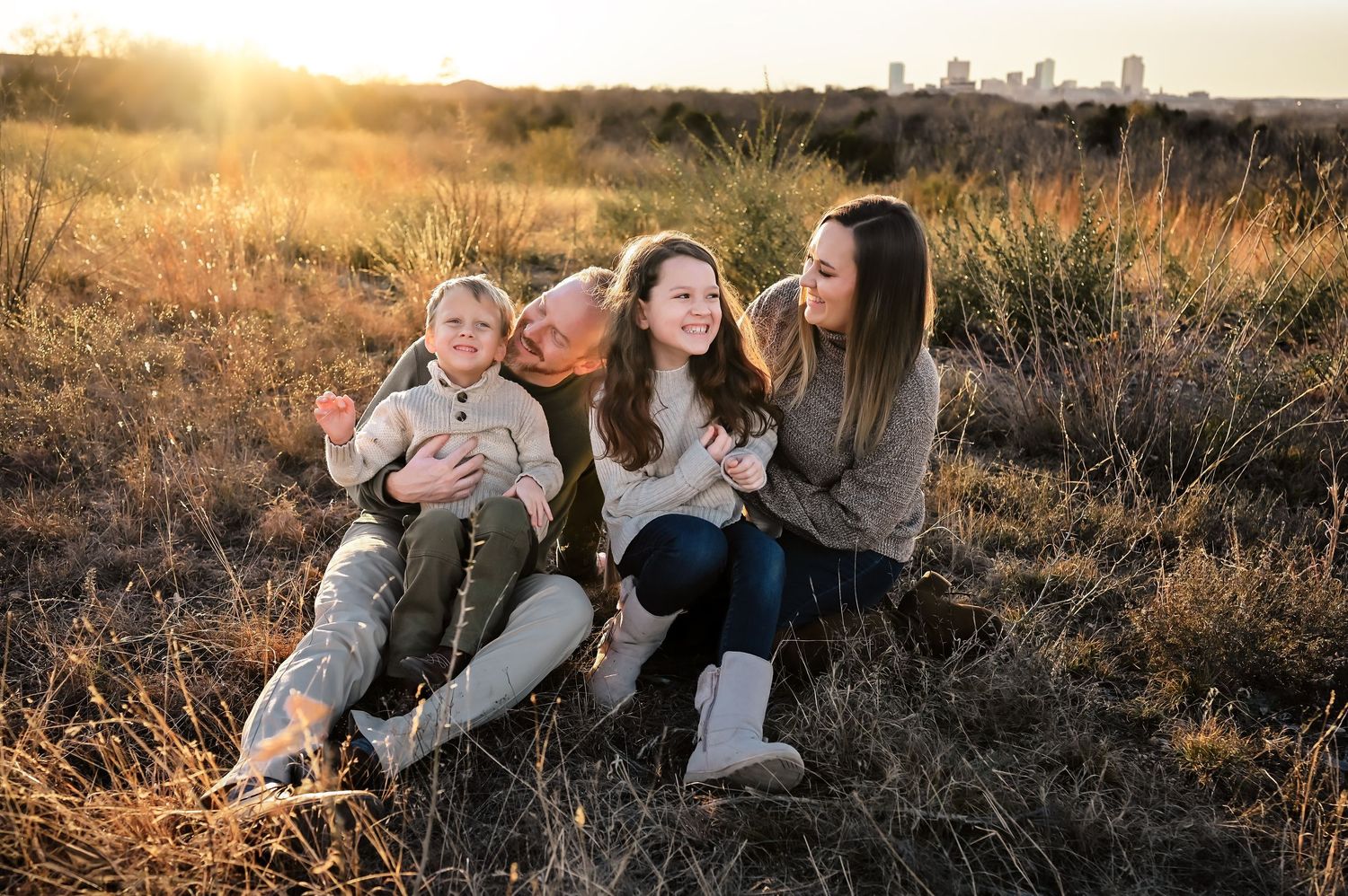 Family of four sitting in golden field at sunset, children laughing with mother and older sibling, city skyline in background.