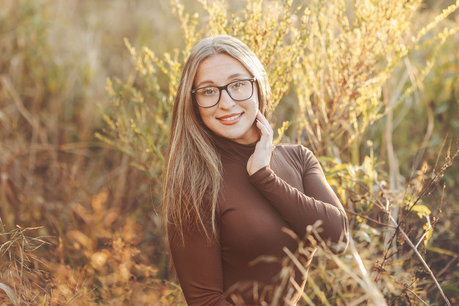 Young blonde woman wearing glasses and brown turtleneck smiling outdoors in golden sunlight among wild plants.