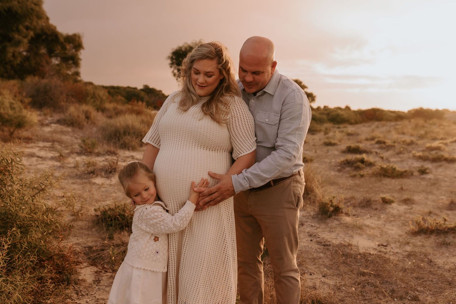 Family of four walking through a sunny golden field, parents holding children's hands in warm afternoon light.