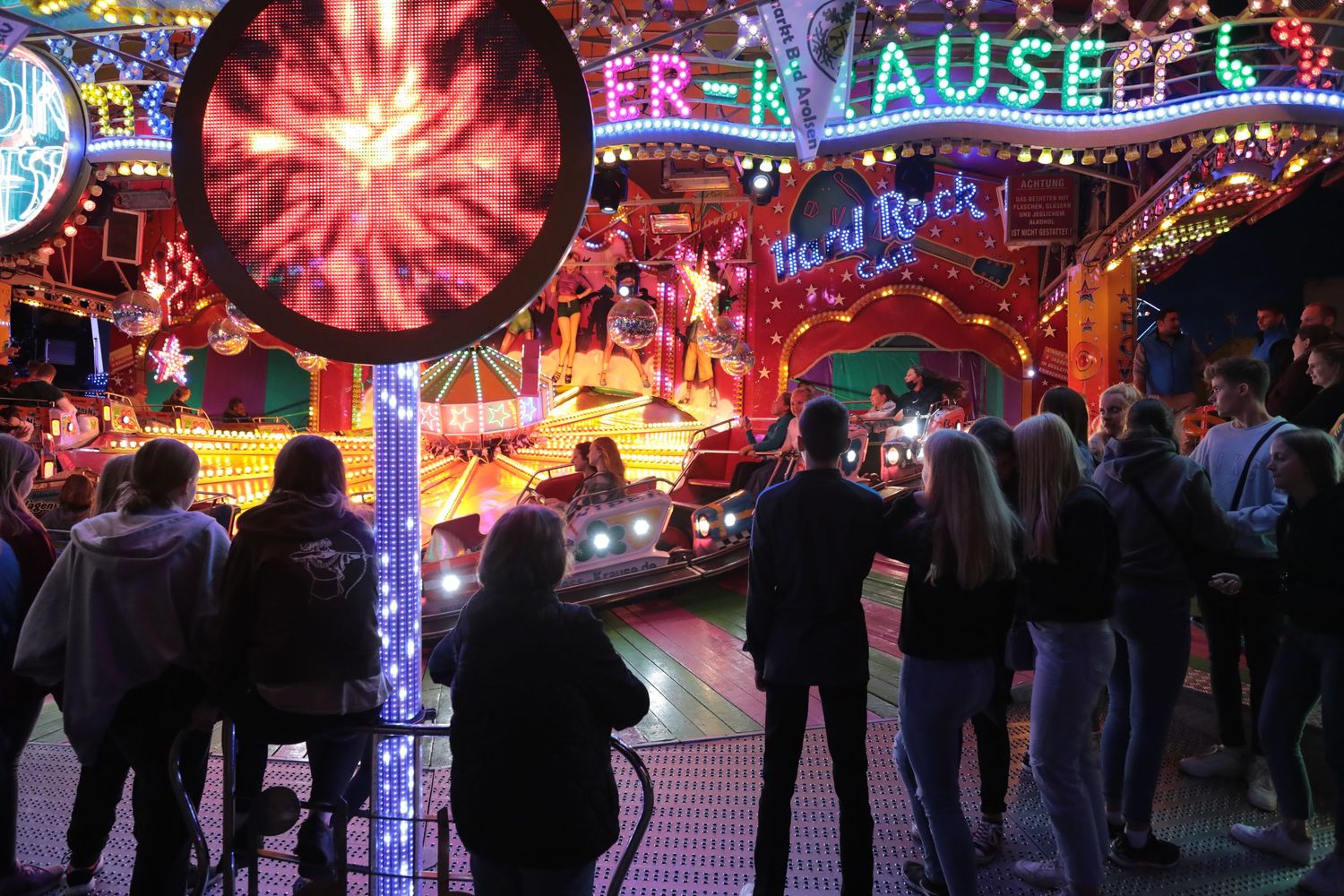 Crowds gather at a vibrant illuminated fairground ride at night with colorful neon lights.