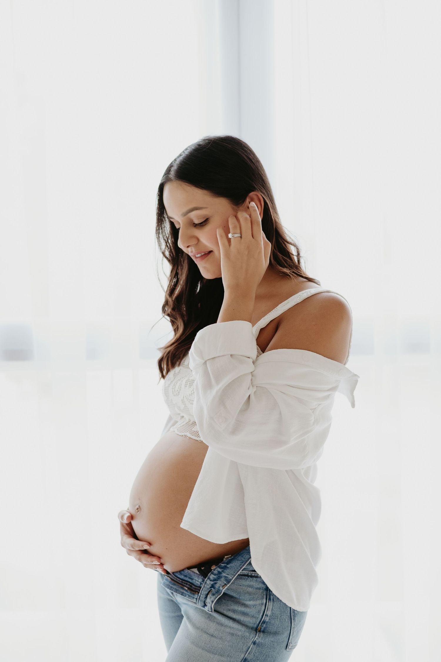 Smiling pregnant woman in white off-shoulder shirt and jeans cradling her baby bump against a bright white background.