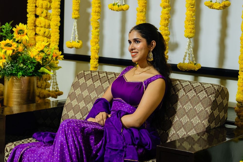 Woman in purple saree sitting on sofa with yellow floral decorations and sunflowers in background.