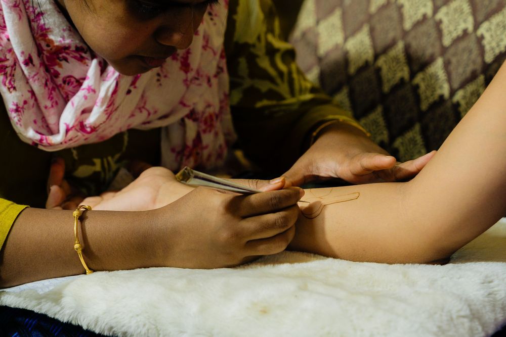Woman applying intricate henna mehndi design on a hand using a cone applicator on a white surface.