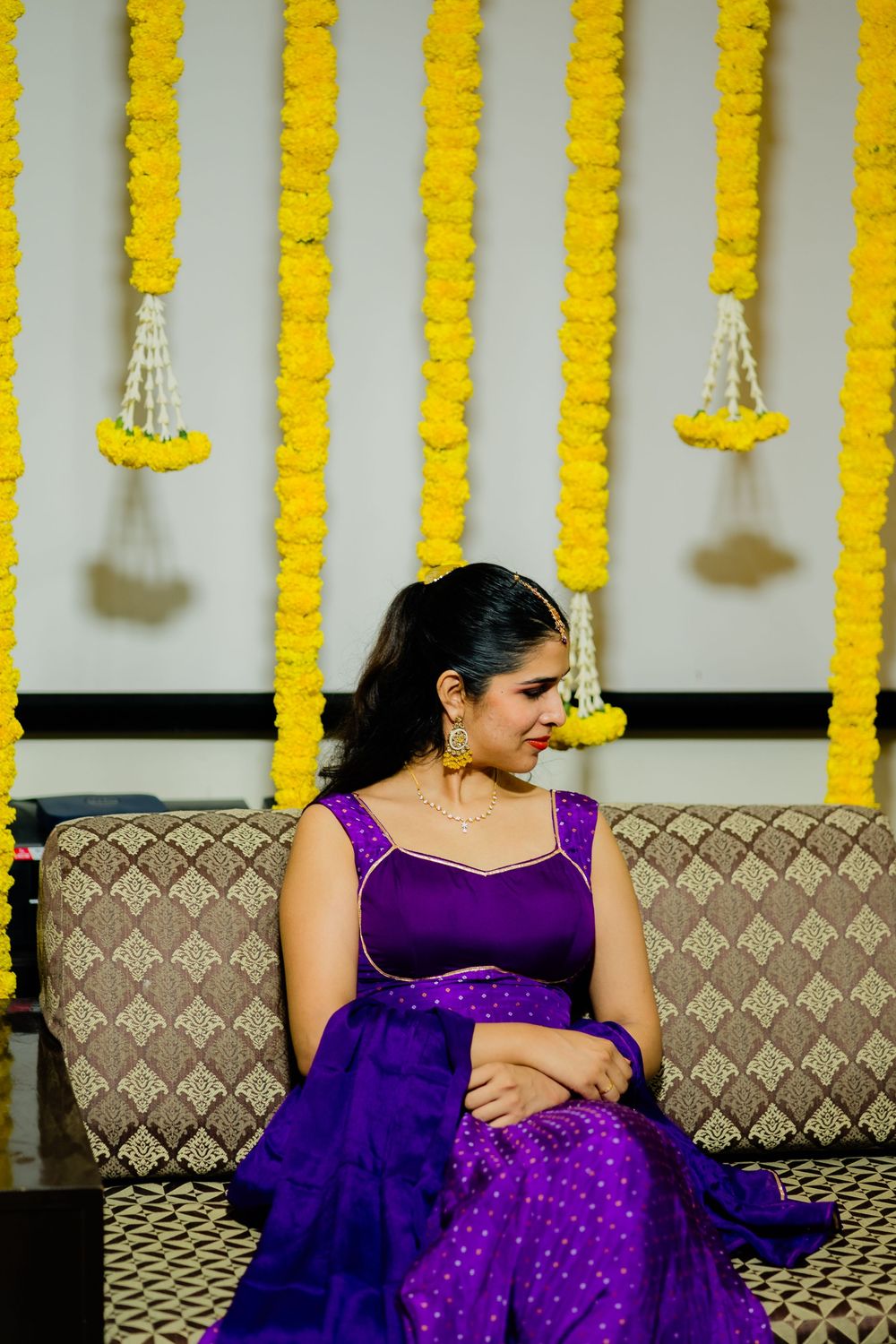 Woman in purple lehenga sitting on sofa with yellow marigold flower garlands backdrop at Indian ceremony.