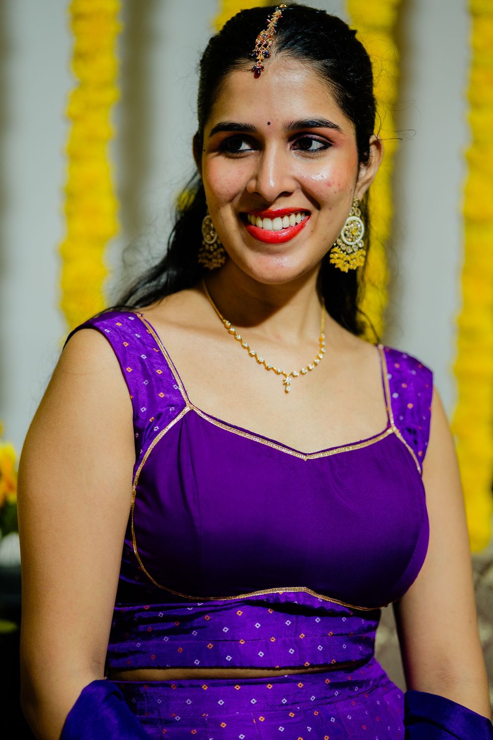Smiling Indian bride in purple lehenga with gold jewelry and maang tikka at mehndi ceremony.