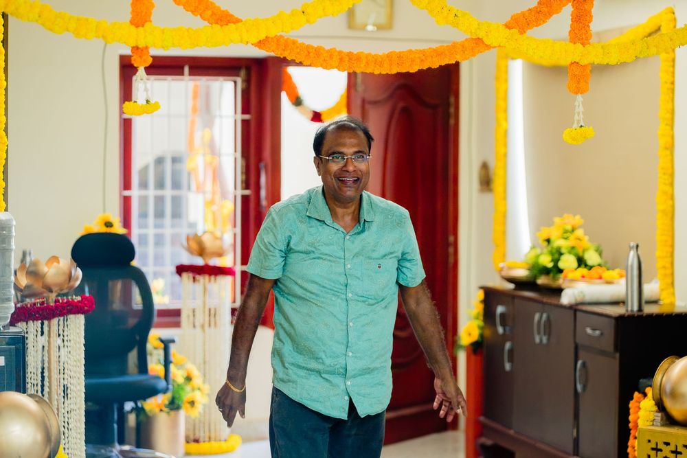 Man in teal shirt smiling indoors decorated with marigold garlands for an Indian celebration.