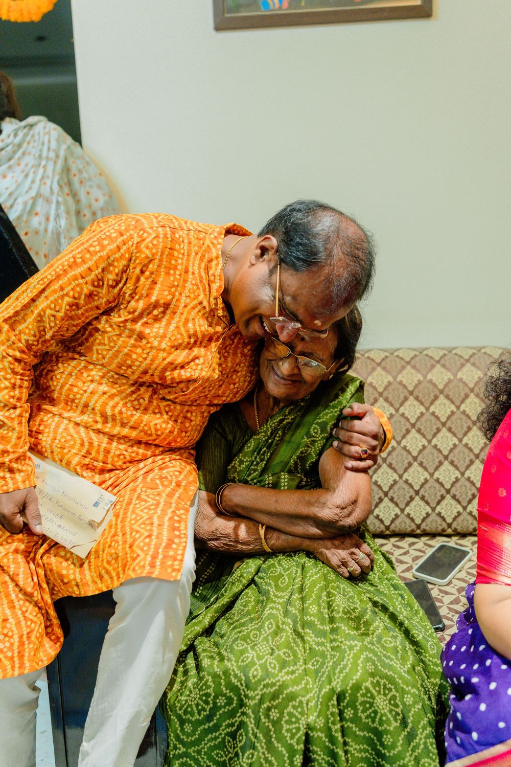 Elderly Indian couple sharing a tender embrace, wearing traditional orange kurta and green saree during a family celebration.