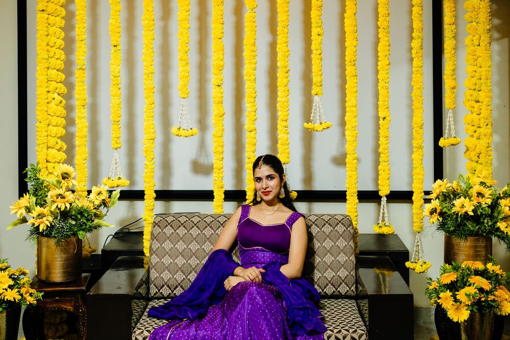 Bride in purple lehenga sits elegantly against vibrant yellow marigold flower backdrop during Indian wedding ceremony.