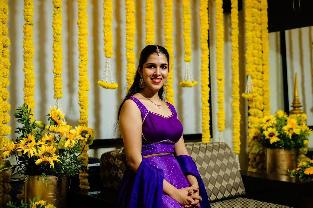 Woman in purple lehenga poses at Indian celebration with yellow marigold garlands and sunflowers backdrop.