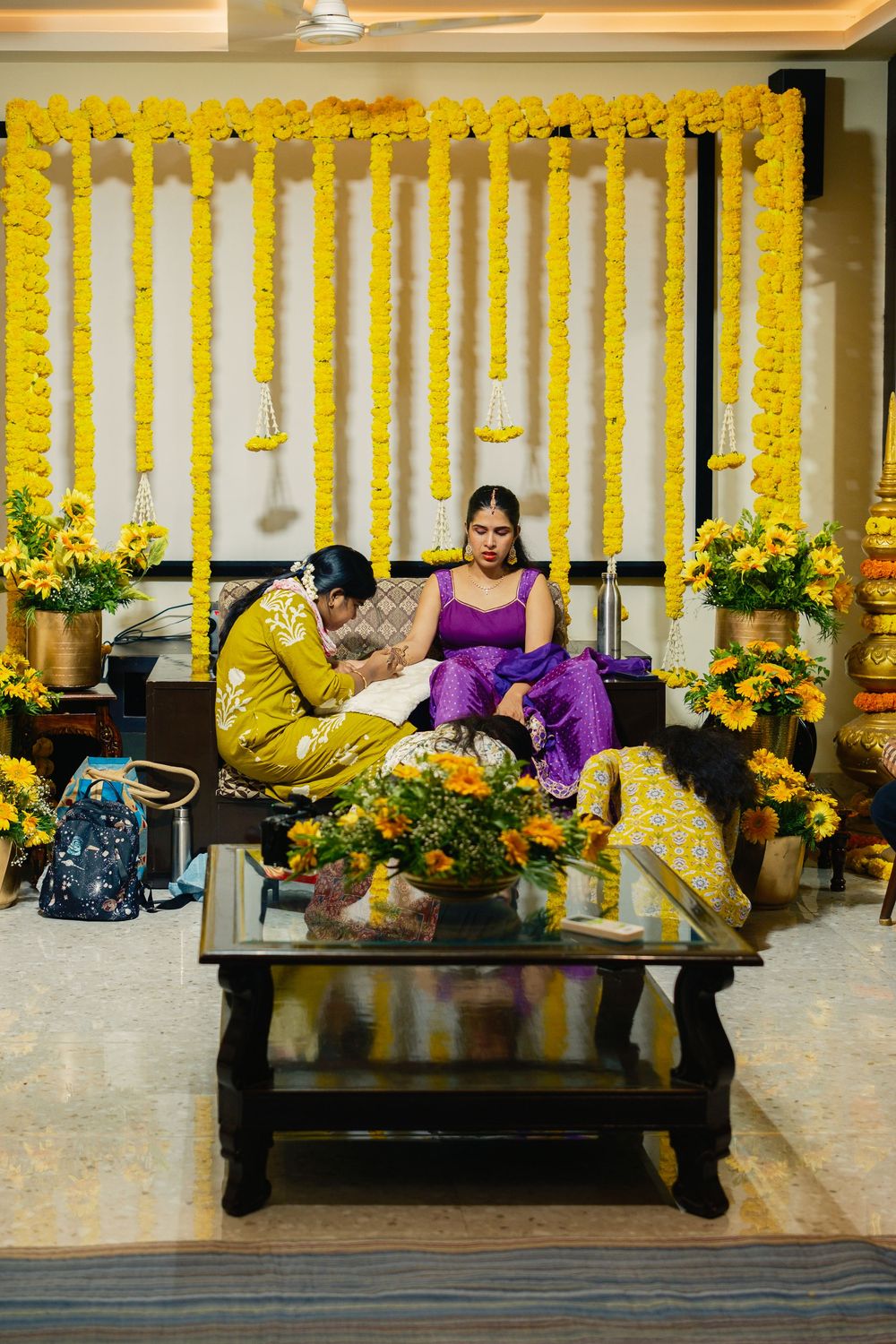 Indian bride in purple sits for mehndi ceremony amid vibrant yellow marigold floral decorations indoors.