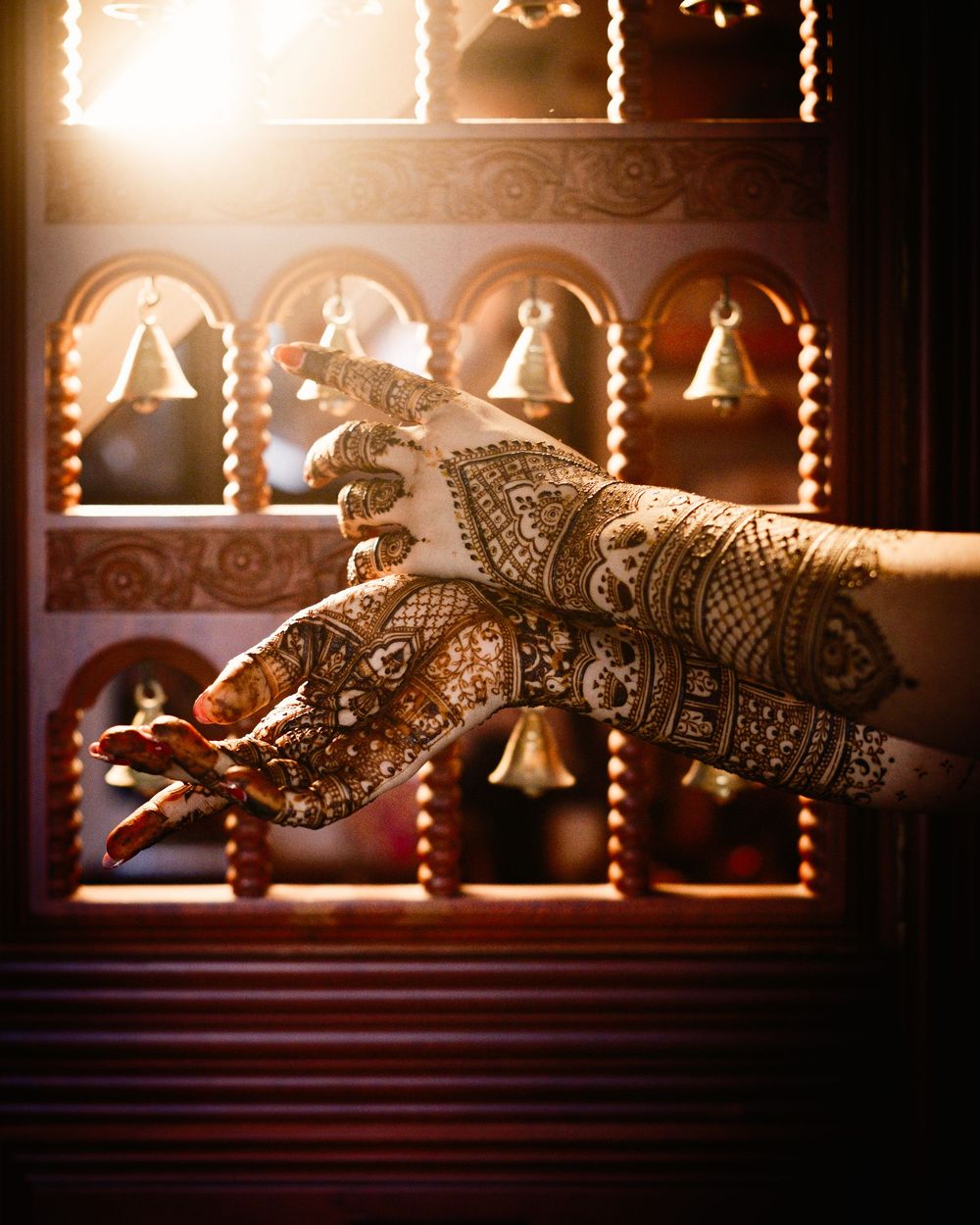 Intricate bridal mehndi henna designs on hands posed near ornate temple bells in warm golden light.