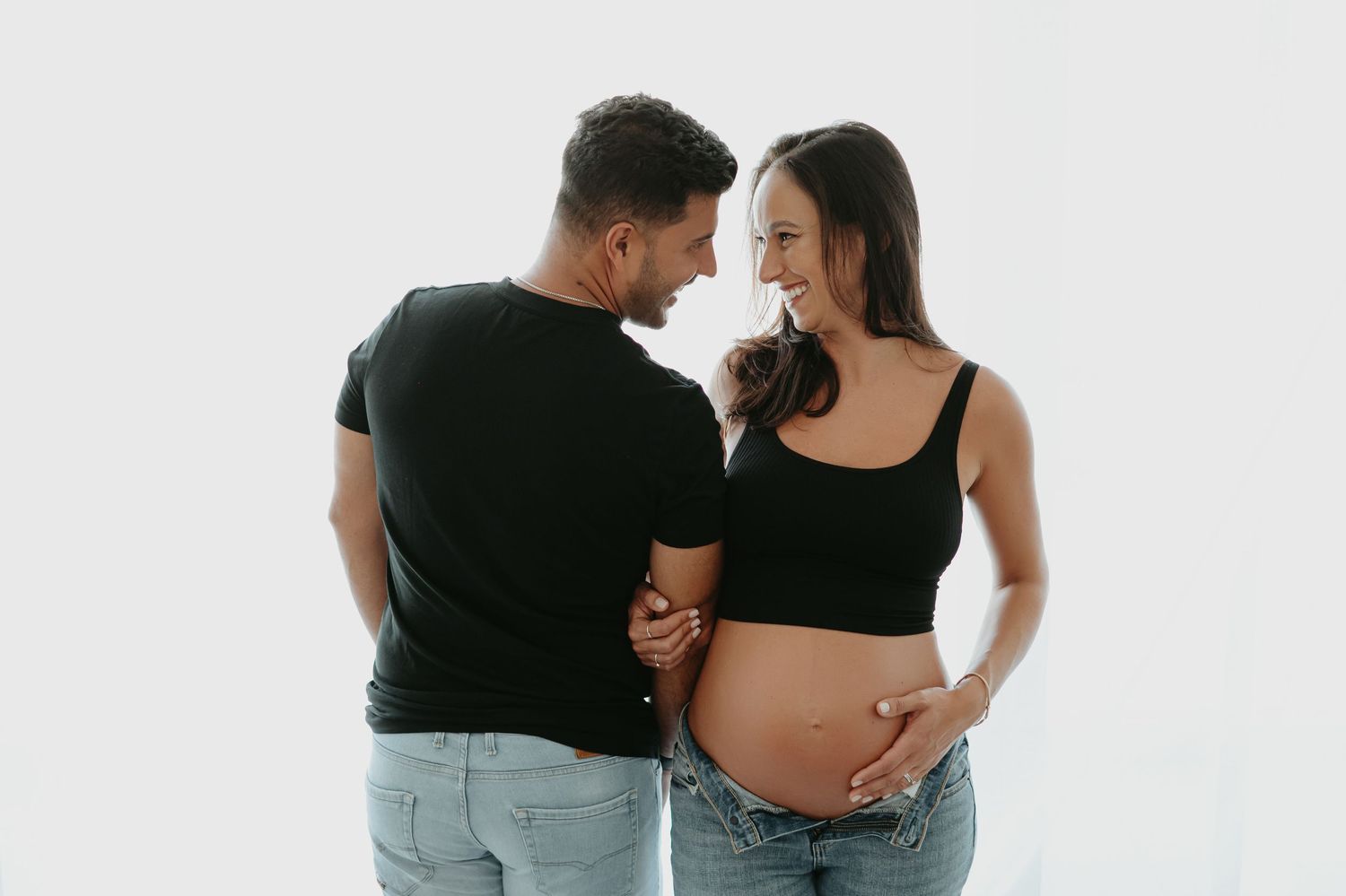 Pregnant woman in black sleeveless bodysuit cradling baby bump, smiling softly against neutral gray background.
