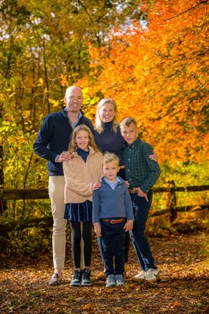 Family of five posing outdoors among vibrant autumn foliage with orange and yellow leaves in background.