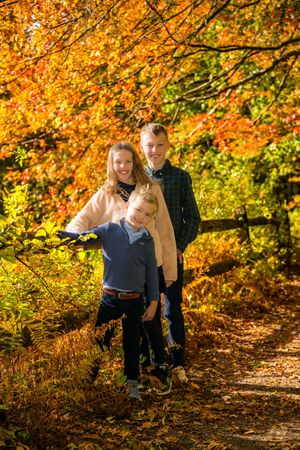 Three children smile together on a leaf-covered path surrounded by vibrant autumn foliage in orange, red, and yellow.