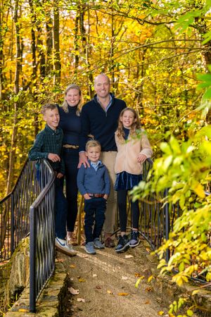 Family of five smiling on a bridge surrounded by vibrant autumn foliage in a forest setting.