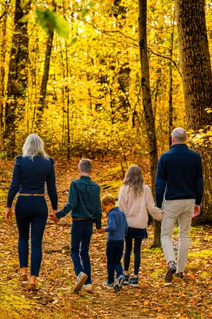 Family of five walking hand-in-hand through a golden autumn forest on a leaf-covered path.