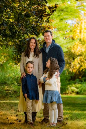 Family of four posing outdoors in autumn garden, parents with two children smiling among golden-green foliage.
