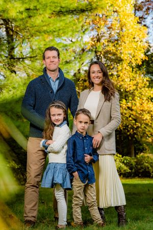 Family of four posing outdoors in autumn garden with colorful yellow and green foliage backdrop.