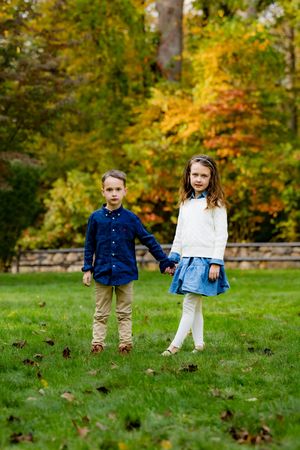 Two young children holding hands on green grass with colorful autumn trees in the background.