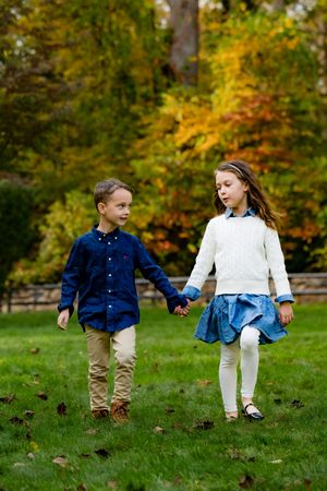 Two children holding hands and walking on green grass with colorful autumn trees in the background.