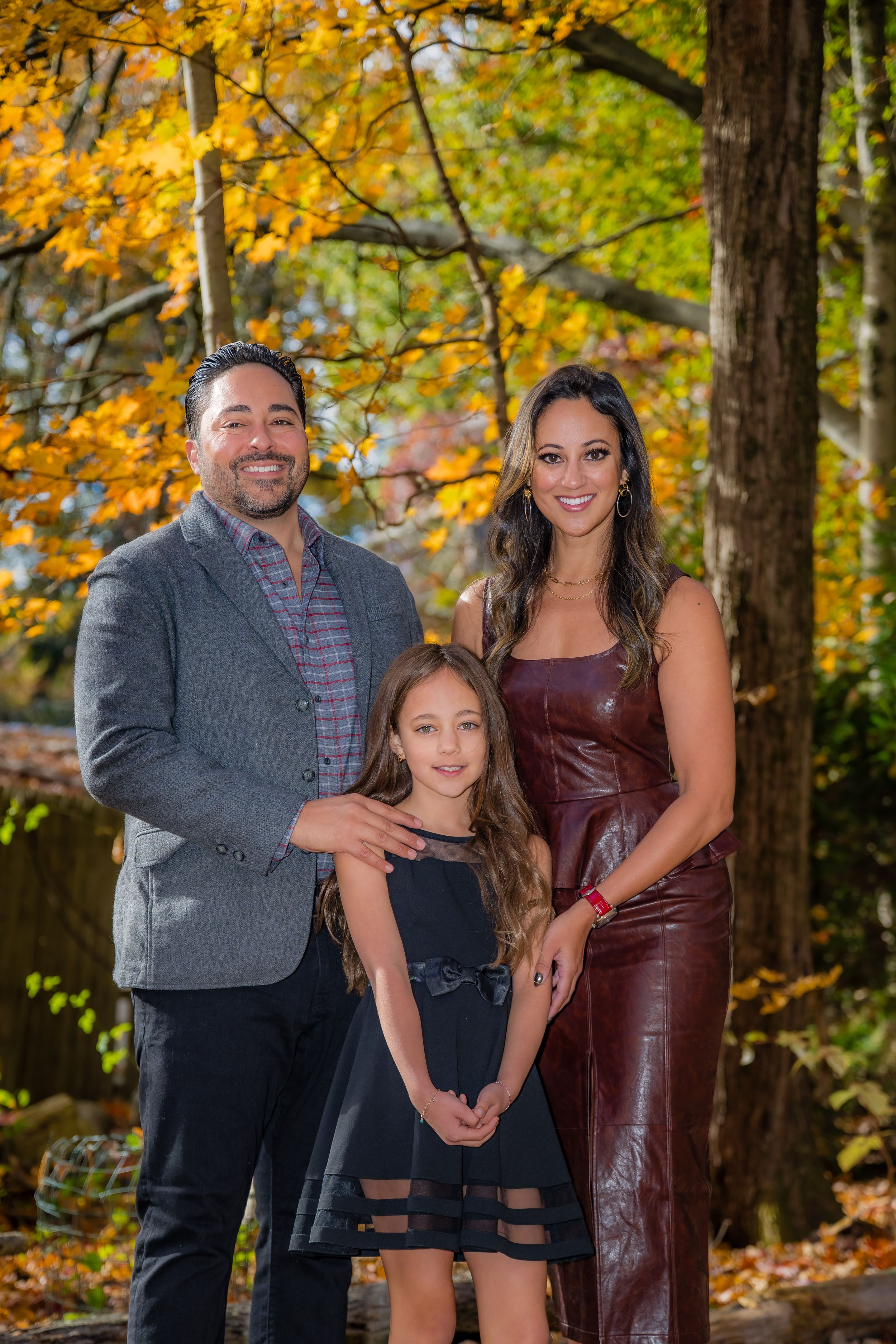 Family of three posing outdoors among vibrant autumn foliage, man in gray blazer, woman in burgundy dress, young girl in black dress.