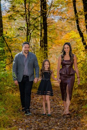 Family of three walking hand-in-hand on a leaf-covered path surrounded by colorful autumn foliage.