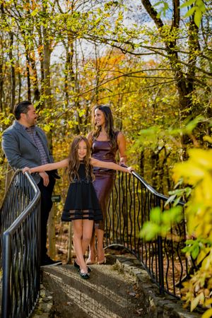 Family of three posing on an outdoor bridge surrounded by colorful autumn foliage, dressed in elegant attire.
