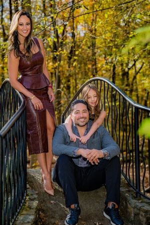 Family of three posing on stone steps surrounded by autumn foliage, woman in burgundy dress, man seated, child behind him.