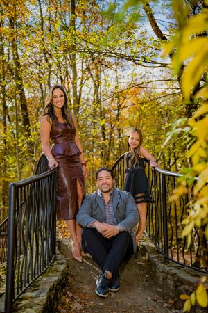 Family of three posing on a stone bridge surrounded by vibrant autumn foliage in a scenic woodland setting.
