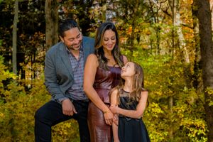 Family of three smiling together in autumn forest, parents holding hands with daughter in fall foliage setting.