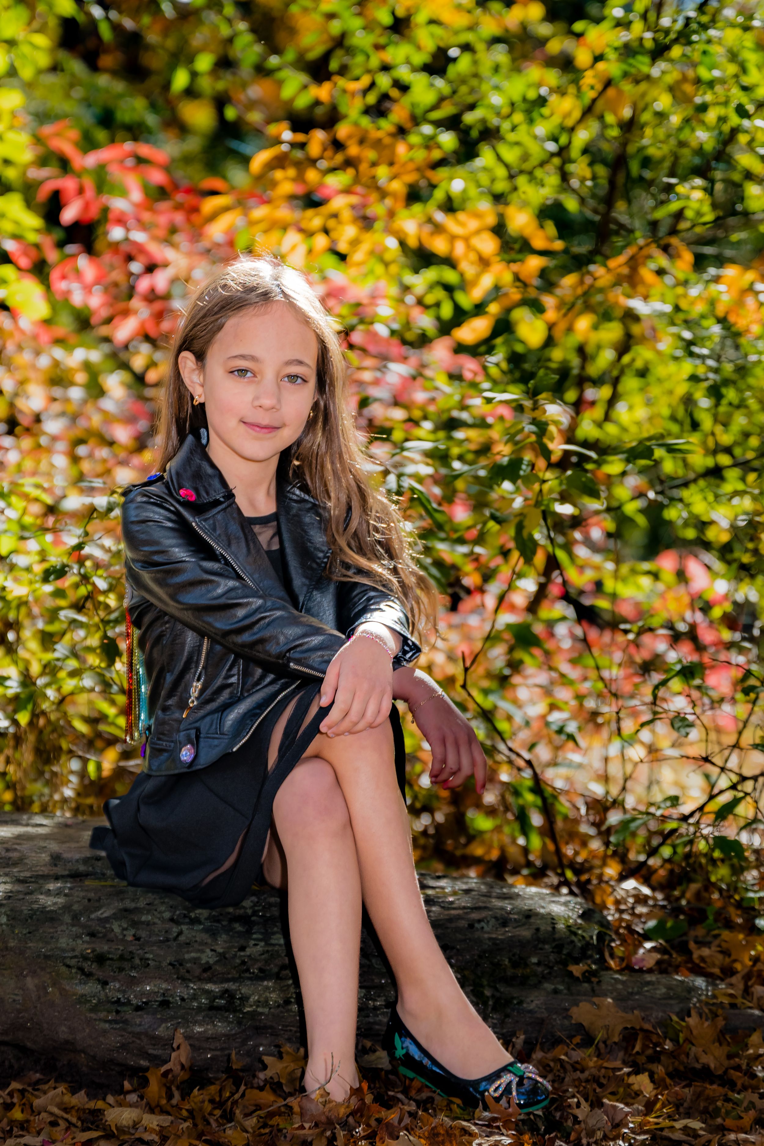 A young girl in a black leather jacket and dress sits on a rock surrounded by colorful autumn foliage.