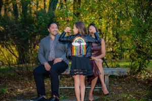 Family of three posing on a park bench in autumn, child wearing a rainbow leather jacket between smiling parents.