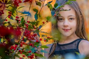 A young girl with long brown hair poses among colorful autumn leaves and red berries.